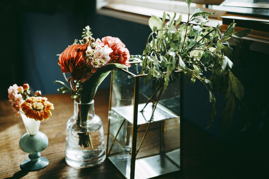 木のテーブルに、赤や橙の花を飾った3つの器と緑。本人のAND PLANTS花瓶記事の写真。
